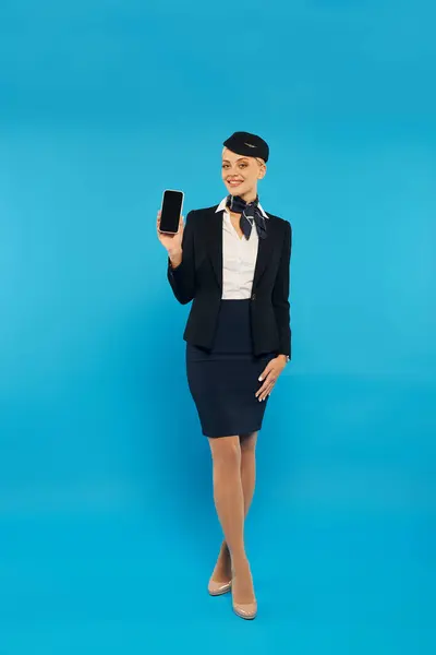 Full length of joyful stewardess in uniform showing smartphone with blank screen on cyan backdrop — Stock Photo