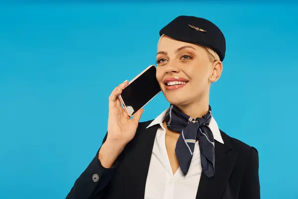 Joven y alegre azafata en elegante uniforme hablando en smartphone con pantalla en blanco en azul - foto de stock