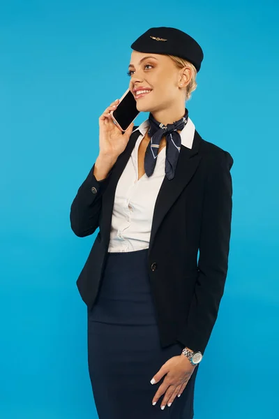 Hostess felice ed elegante con borsa da viaggio che parla sul cellulare su sfondo blu, lunghezza intera — Foto stock