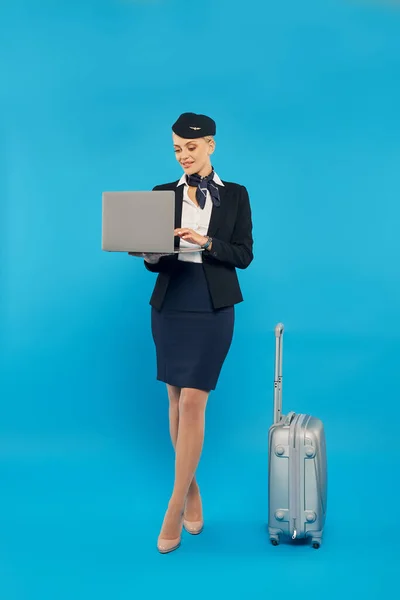 Piena lunghezza di affascinante hostess aerea in uniforme utilizzando il computer portatile vicino alla valigia su blu, piena lunghezza — Foto stock