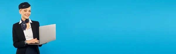 Aeromoça positiva em laptop uniforme elegante segurando e sorrindo para a câmera em azul, banner — Fotografia de Stock