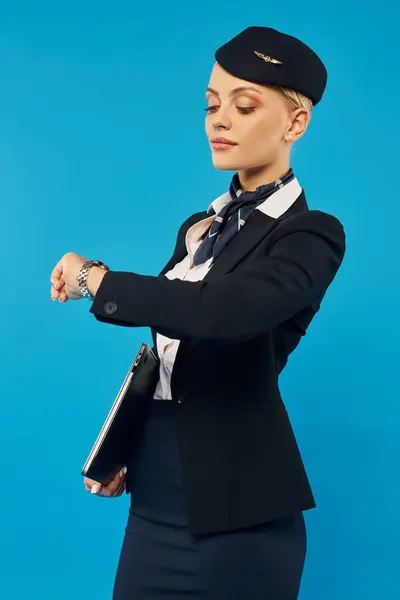 Jeune hôtesse en uniforme élégant regardant montre-bracelet tout en vérifiant l'heure sur fond bleu — Photo de stock