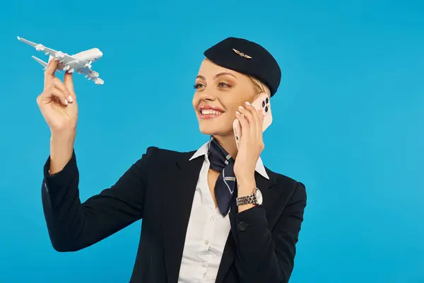Azafata feliz en uniforme sosteniendo modelo de avión y hablando om teléfono inteligente sobre fondo azul - foto de stock