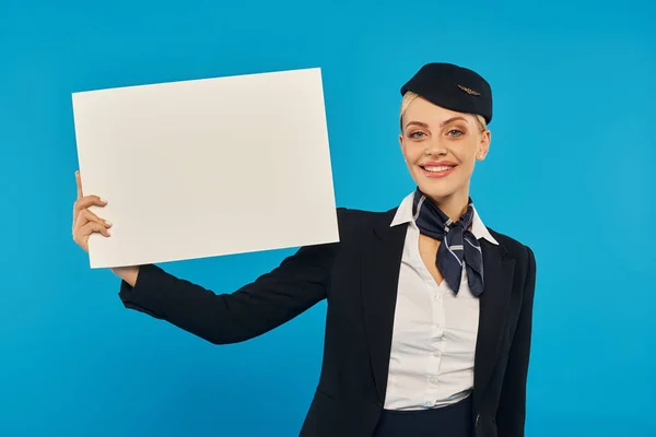 Young cheerful stewardess in elegant uniform standing with empty poster on blue studio backdrop — Stock Photo