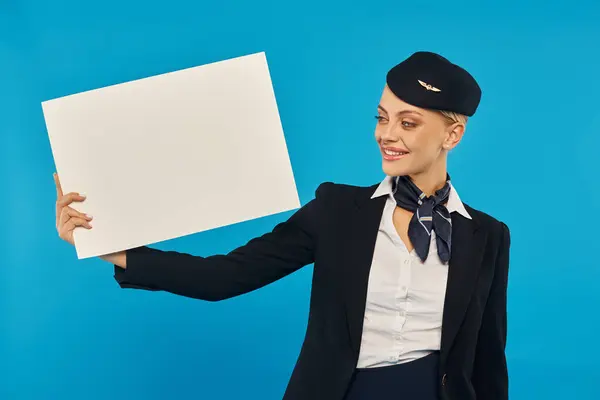 Усміхнена повітряна господиня в уніформі авіакомпаній тримає порожній плакат, стоячи на синьому фоні — стокове фото