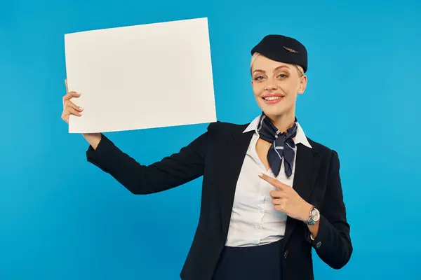 Azafata elegante y sonriente en uniforme señalando con el dedo en el cartel en blanco sobre fondo azul - foto de stock