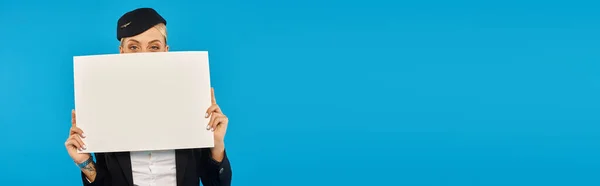 Young stewardess in corporate uniform obscuring face with blank placard on blue background, banner — Stock Photo