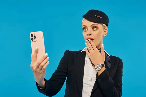 Surprised and impressed stewardess looking at mobile phone and covering mouth with hand on blue — Stock Photo
