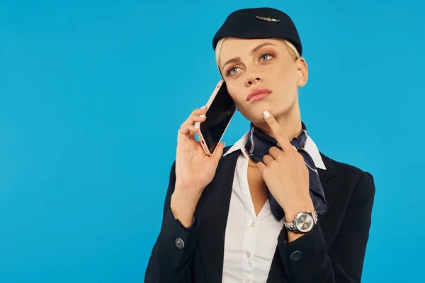 Hôtesse de l'air réfléchie en uniforme parlant sur le téléphone mobile et regardant loin sur fond bleu — Photo de stock