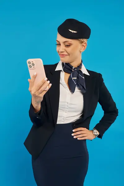 Anfitriona del aire encantado en uniforme mirando el teléfono inteligente y de pie con la mano en la cadera en azul - foto de stock