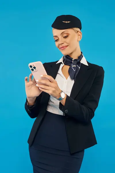 Aeromoça sorridente em mensagens uniformes elegantes no telefone celular enquanto está de pé no fundo azul — Fotografia de Stock