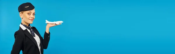 Hôtesse élégante heureuse dans unform tenant modèle d'avion et souriant à la caméra sur bleu, bannière — Photo de stock