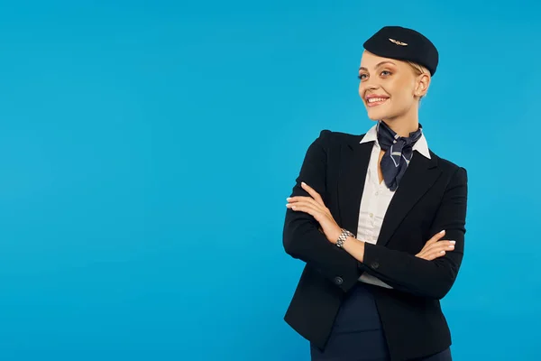 Joyful air hostess in corporate uniform posing with folded arms on blue backdrop, airline industry — Stock Photo