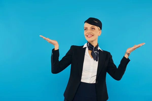 Young and overjoyed stewardess in elegant uniform looking up and showing wow gesture on blue — Stock Photo