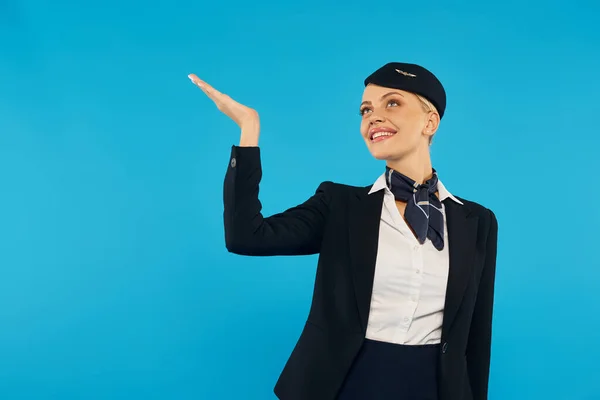 Excited and elegant stewardess in corporate uniform looking up and pointing with hand on blue — Stock Photo