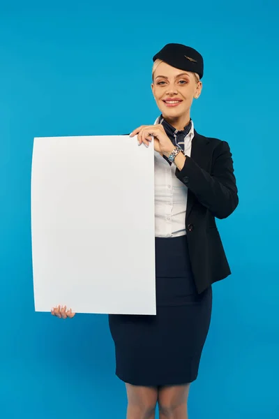 Alegre azafata de aire en elegante uniforme sosteniendo pancarta en blanco y sonriendo a la cámara sobre fondo azul - foto de stock