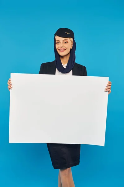 Azafata feliz en uniforme de las aerolíneas árabes sosteniendo pancarta en blanco sobre fondo de estudio azul - foto de stock