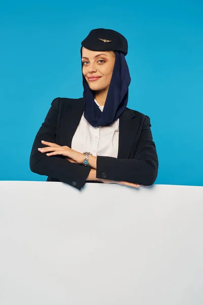 Stewardess der arabischen Fluggesellschaften mit verschränkten Armen lächelt in die Kamera neben einem leeren Plakat auf blauem Hintergrund — Stockfoto