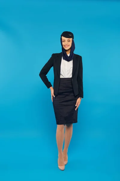 Jeune femme dans les compagnies aériennes arabes uniforme et foulard posant avec la main sur la hanche sur bleu, pleine longueur — Photo de stock