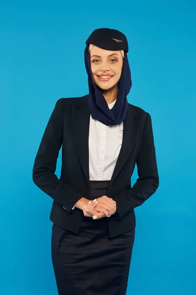 Happy stewardess in uniform and headscarf smiling at camera on blue, arabian airlines dress code — Stock Photo