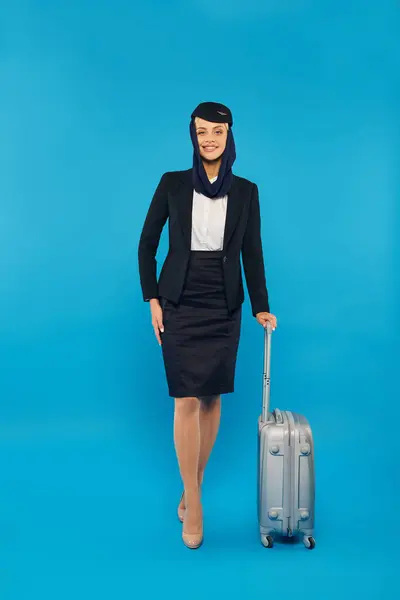 Elegant stewardess in uniform of arabian airlines standing with hand on hip and travel bag on blue — Stock Photo