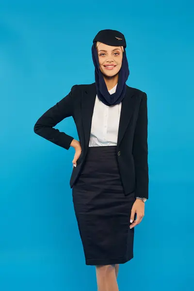 Jolly air hostess in uniform and headscarf posing with hand on hip on blue, arabian airlines — Stock Photo