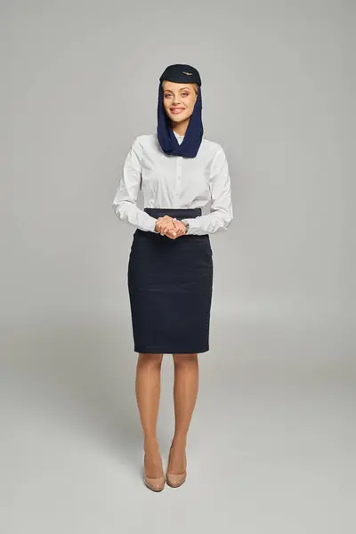 Full length of happy air hostess in arabian airlines uniform standing and looking at camera on grey — Stock Photo