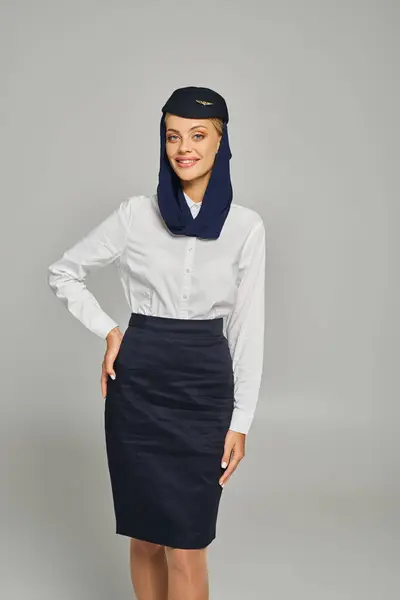 Stylische und gut gelaunte Stewardess in Uniform arabischer Airlines posiert mit der Hand an der Hüfte auf grau — Stockfoto