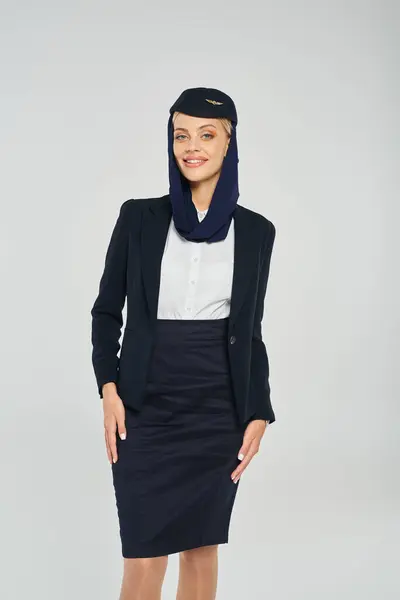 Hôtesse de l'air souriante en foulard et uniforme d'entreprise des compagnies aériennes arabes regardant la caméra sur gris — Photo de stock