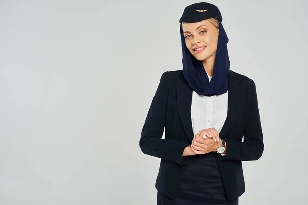 Joyous air hostess in headscarf and elegant uniform of arabian airlines smiling at camera on grey — Stock Photo