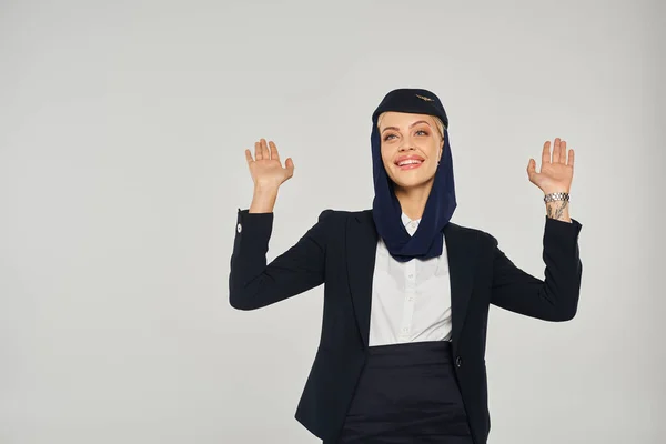 Aufgeregte Stewardess in Uniform arabischer Fluggesellschaften blickt auf und winkt vor grauem Hintergrund — Stockfoto