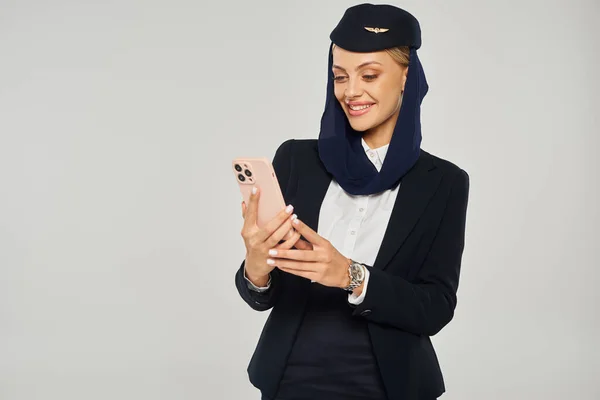 Lächelnde Stewardess in Uniform arabischer Fluggesellschaften, die auf dem Handy vor grauem Hintergrund Nachrichten sendet — Stockfoto