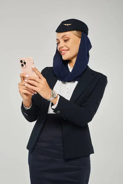 Gut gelaunte Stewardess in Uniform arabischer Fluggesellschaften vernetzt sich auf Handy vor grauem Hintergrund — Stockfoto