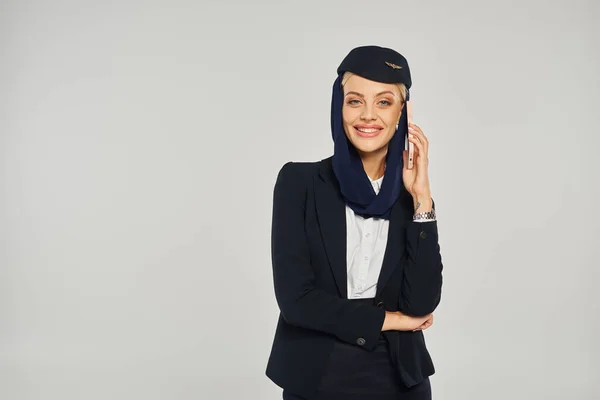Graziöse Stewardess arabischer Fluggesellschaften, die am Handy telefoniert und in grau in die Kamera lächelt — Stockfoto