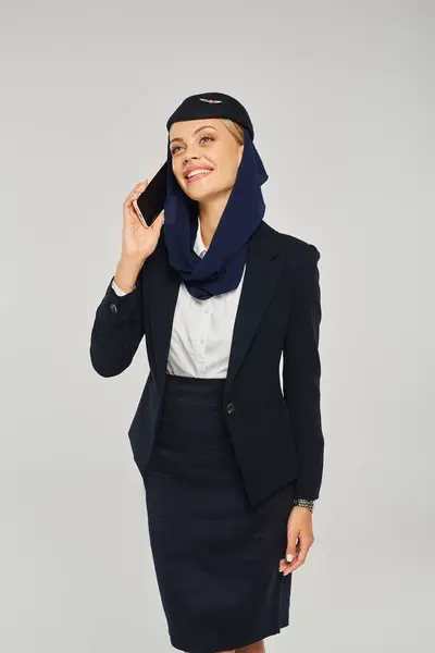 Hôtesse de l'air souriante dans l'uniforme des compagnies aériennes arabes parler sur smartphone et détourner les yeux sur gris — Photo de stock