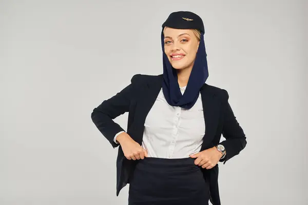 Sorridente arábica aeromoça linhas aéreas em uniforme e lenço de cabeça posando com as mãos na cintura em cinza — Fotografia de Stock