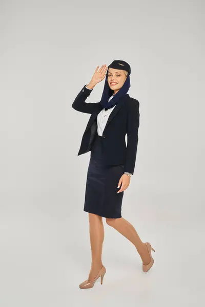 Elegante azafata de las aerolíneas árabes en uniforme caminar y agitar la mano en gris, longitud completa - foto de stock