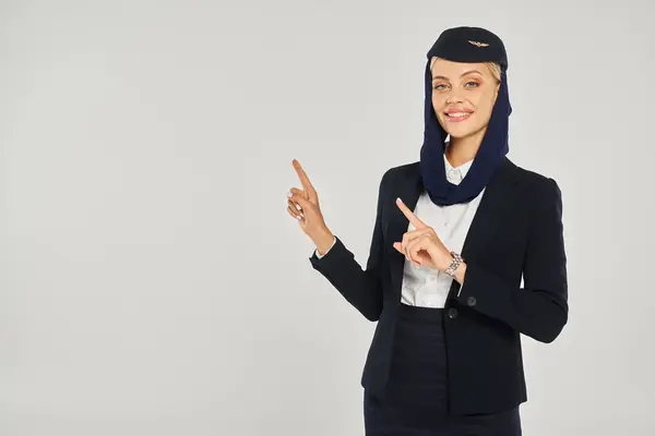Charmante Stewardess in Uniform arabischer Fluggesellschaften lächelt in die Kamera und zeigt mit den Fingern auf grau — Stockfoto