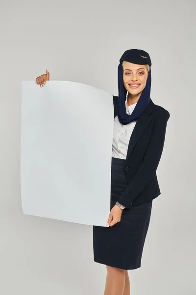 Happy arabian airlines stewardess in elegant uniform holding empty poster on grey backdrop — Fotografia de Stock