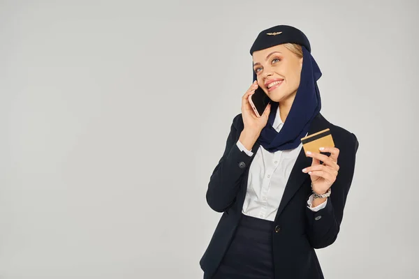 Счастливая стюардесса арабских авиалиний в форме с кредитной картой разговаривает на смартфоне на сером — стоковое фото
