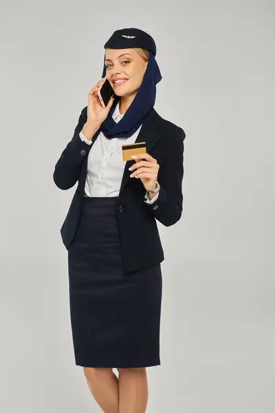 Счастливая стюардесса в униформе арабских авиалиний держит кредитную карту и разговаривает по смартфону на сером — стоковое фото