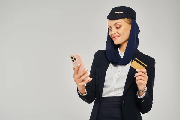Lächelnde Stewardess in Uniform arabischer Fluggesellschaften, die Kreditkarte in der Hand hält und grau auf ihr Smartphone blickt — Stockfoto