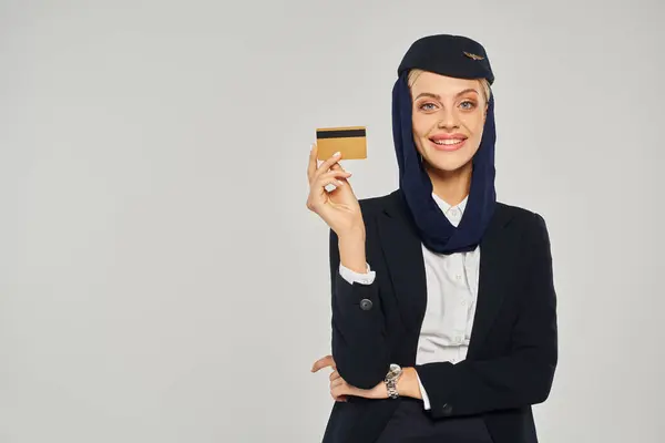 Young and elegant arabian airlines stewardess with credit card smiling at camera on grey backdrop — Stock Photo
