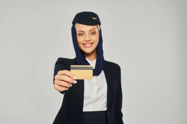 Junge und gut gelaunte arabische Stewardess in Uniform zeigt Kreditkarte vor Kamera auf grau — Stockfoto