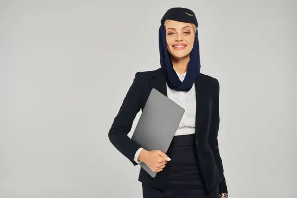 Positive arabian airlines stewardess in uniform with laptop smiling at camera on grey backdrop — Stock Photo