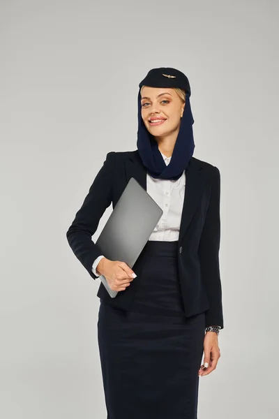 Encantadora y elegante azafata de las aerolíneas árabes sosteniendo el ordenador portátil y sonriendo a la cámara en gris - foto de stock