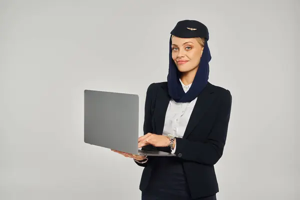 Hôtesse de l'air des compagnies aériennes arabes heureux en uniforme élégant tenant ordinateur portable tout en se tenant sur gris — Photo de stock
