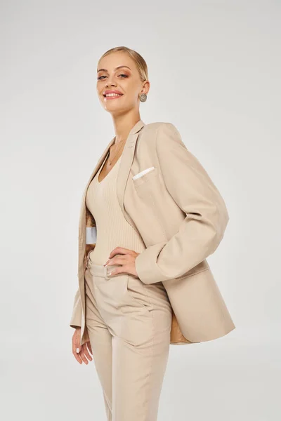 Selbstbewusste und charmante Frau im beigen Anzug posiert mit der Hand an der Hüfte auf grauer, moderner Mode — Stockfoto