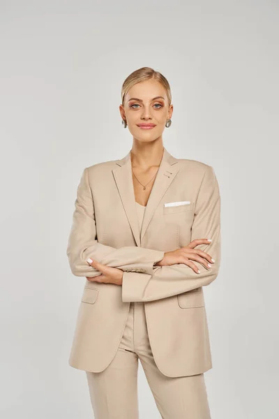 Lächelnde Frau im beigen Anzug posiert mit verschränkten Armen und blickt in die Kamera auf grau, Modetrend — Stockfoto