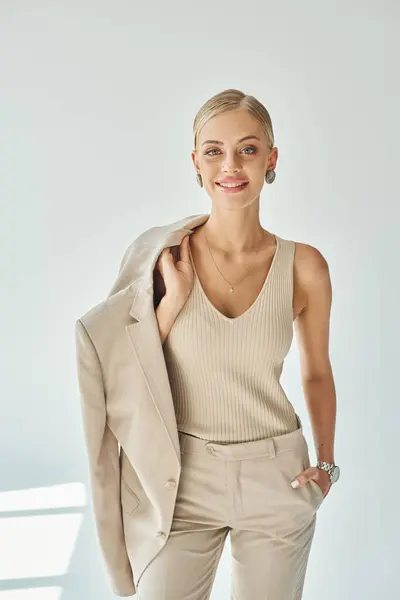 Donna sorridente in abiti beige che tiene la giacca mentre tiene la mano in tasca su grigio, bellezza e stile — Foto stock